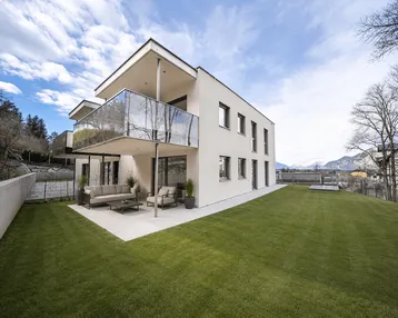 Moderne Wohnanlage mit großzügiger Terrasse, Rasenfläche und Bergblick unter blauem Himmel.