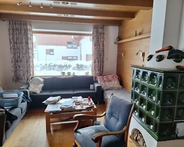 Rustikales Wohnzimmer mit Kachelofen, Holzbalkendecke und großem Fenster mit Winterblick.