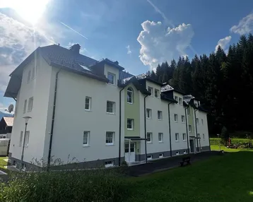 Mehrfamilienhaus mit weißer und grüner Fassade, umgeben von Bäumen und Grünflächen unter blauem Himmel.