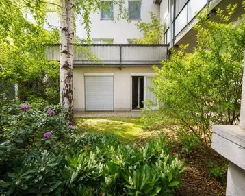 Grüner Gartenbereich mit Rasenfläche und Zugang zum Gebäude, umgeben von Bäumen und Sträuchern.