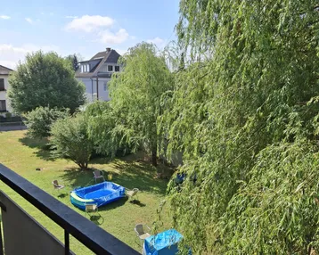 Blick vom Balkon auf den Gemeinschaftsgarten mit Bäumen und einem kleinen Planschbecken unter blauem Himmel.