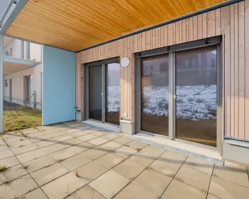 Gepflasterte Terrasse mit Holzwand und Überdachung, angrenzend an eine Grünfläche.