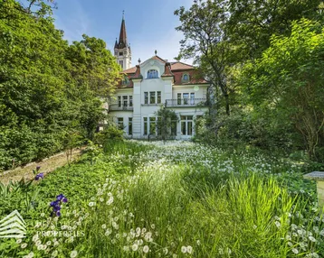 Historische Villa mit weitläufigem Garten und Blick auf die Kirchturmspitze, umgeben von üppigem Grün.