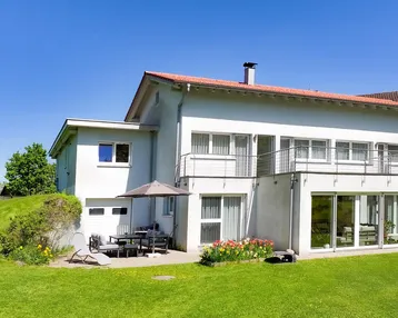 Weißes Einfamilienhaus mit rotem Dach, großem Garten und möblierter Terrasse unter blauem Himmel.