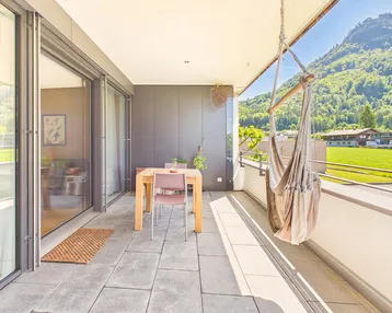 Geräumiger Balkon mit Fliesenboden, Esstisch und Hängesessel, umgeben von grünen Bergen.