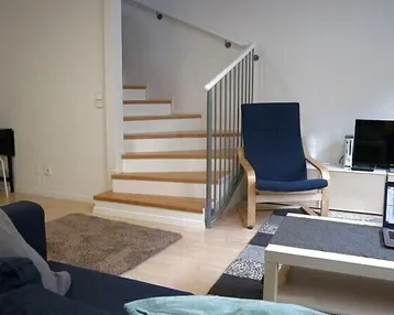Gemütliches Wohnzimmer mit Sofa, Fernseher und Zugang zu einer Holztreppe im Hintergrund.