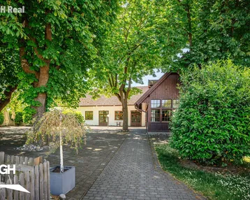 Ein charmantes Gebäude mit traditioneller Architektur, umgeben von üppigem Grün und hohen Bäumen, mit einem gepflasterten Weg zum Eingang.