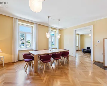 Geräumiges Esszimmer mit Fischgrätparkett, hohen Fenstern und Zugang zu einem weiteren Raum.