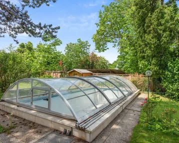 Überdachter Swimmingpool im Gartenbereich, umgeben von üppigem Grün und einem kleinen Gartenhaus.