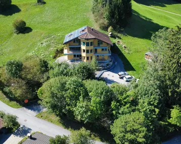 Luftaufnahme eines gelben Hauses mit Solaranlage auf dem Dach, eingebettet in grüne Hügel und Bäume.
