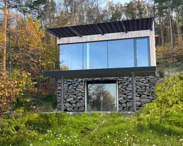 Modernes Haus mit großer Glasfassade, Steinsockel und Solarpaneelen auf dem Dach, umgeben von Bäumen.
