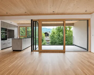 Heller offener Wohnbereich mit Holzboden, Holzdecke und integrierter Küche mit Zugang zum Balkon.