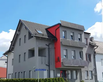 Modernes Mehrfamilienhaus mit grauer Fassade, roten Akzenten und integrierten Solarpaneelen auf dem Dach.