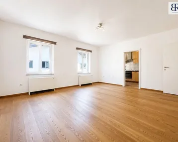 Helles Zimmer mit Holzboden und Blick in die Küche, ideal als Wohn- oder Essbereich.