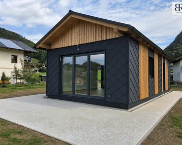 Ansicht des modernen Hauses mit dunkler Fassade, großen Fenstern und Holzakzenten vor bergiger Kulisse.