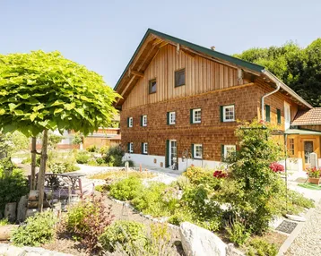 Traditionelles Holzhaus mit gepflegtem Garten und Sitzbereich unter einem Baum an einem sonnigen Tag.