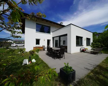 Moderne Hausfassade mit großer gepflasterter Terrasse, Garten und Essbereich im Freien unter blauem Himmel.