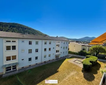 Mehrfamilienhaus mit grüner Rasenfläche und Bergblick unter blauem Himmel.