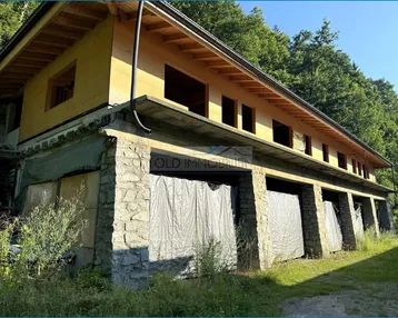 Außenansicht eines im Bau befindlichen Gebäudes mit Steinfundament und Holzkonstruktion, umgeben von Bäumen.