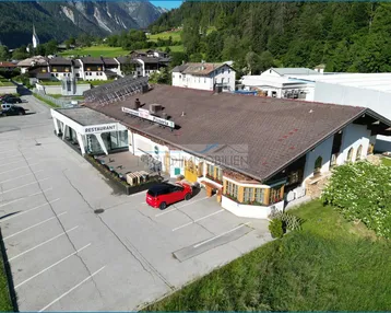 Luftaufnahme eines großen Gebäudekomplexes mit Restaurant und Pizzeria, umgeben von Bergen und einem weitläufigen Parkplatz.