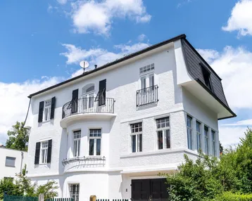 Weißes Mehrfamilienhaus mit Mansarddach, schwarzen Fensterläden und kleinen Balkonen unter blauem Himmel.