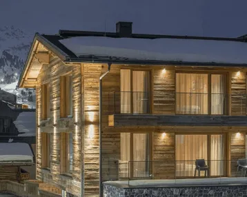 Beleuchtete Holzfassade eines modernen Chalets in verschneiter Berglandschaft bei Nacht.