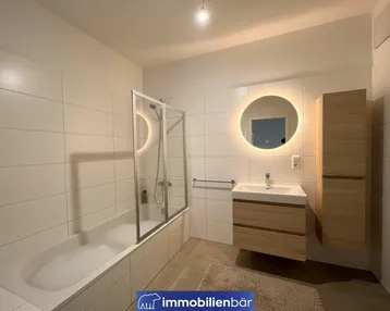 Modernes Badezimmer mit Badewanne, Duschabtrennung und beleuchtetem Spiegel über dem Waschbecken.