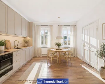 Helle, moderne Wohnküche mit beigefarbenen Schränken, Holzboden und Essbereich am Fenster.
