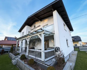 Weißes Einfamilienhaus mit überdachter Terrasse und kleinem Vorgarten unter blauem Himmel.