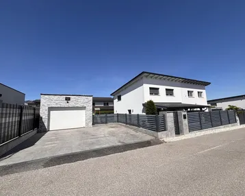 Modernes weißes Einfamilienhaus mit separater Garage, gepflasterter Einfahrt und Zaun.