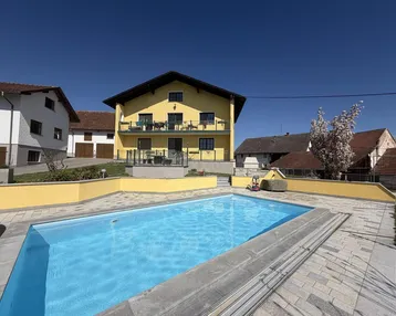 Gelbes Haus mit großem Außenpool und gepflasterter Terrasse unter blauem Himmel.