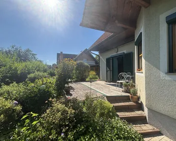 Sonnige Terrasse mit Gartenmöbeln, umgeben von üppigem Grün und Pflanzen.