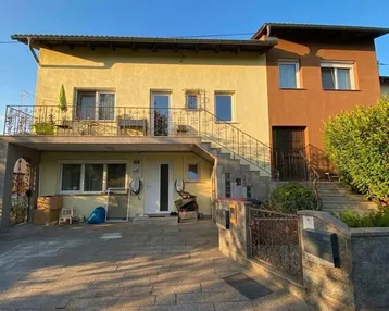Zweifamilienhaus mit gelber Fassade, Balkon und gepflasterter Auffahrt im Sonnenlicht.