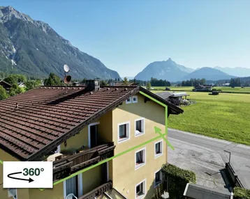Gelbes Gebäude mit braunem Dach in alpiner Landschaft mit Bergblick und grünen Wiesen.