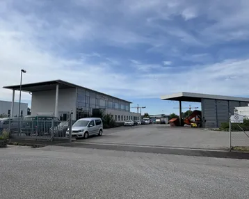 Ansicht des modernen Bürogebäudes mit großem Parkplatz und umliegendem Industriegelände unter bewölktem Himmel.