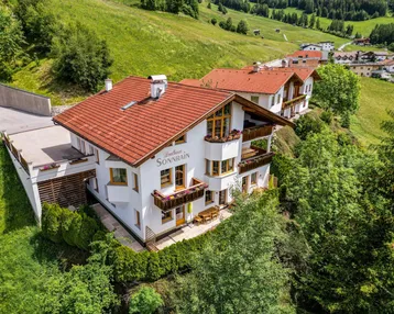 Traditionelles Alpenhaus mit rotem Ziegeldach und mehreren Balkonen, eingebettet in eine grüne Berglandschaft.