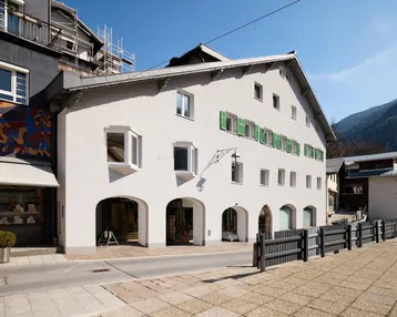 Historisches Gebäude mit weißer Fassade, grünen Fensterläden und Bergblick in einer malerischen Gasse.