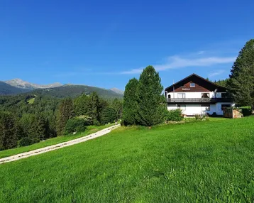 Haus auf einem grünen Hügel mit weitem Blick auf Wälder und Berge unter blauem Himmel.