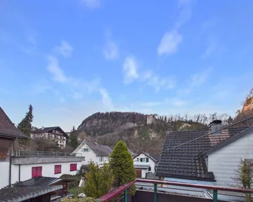 Weitblick über die Dächer der Stadt und bewaldete Hügel mit Burgruine.