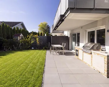 Große gepflasterte Terrasse mit Outdoor-Küche und angrenzendem Rasenbereich, ideal für Entspannung.