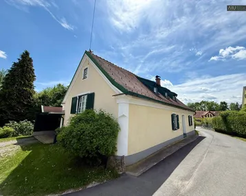 Traditionelles Landhaus mit gelber Fassade, grünen Fensterläden und Satteldach unter blauem Himmel.