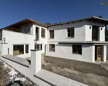 Weiß verputztes Haus mit Holzfenstern und unfertigem Außenbereich unter blauem Himmel.