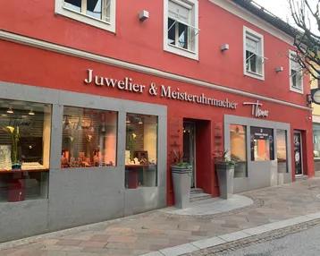 Rote Geschäftsfassade eines Juweliers mit großen Schaufenstern, Eingang und dekorativen Pflanzen am Bürgersteig.