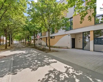 Moderne Wohngebäude-Fassade mit großen Fenstern und begrüntem Gehweg in einer ruhigen Wohngegend.