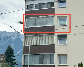 Mehrstöckiges Wohngebäude mit Balkonen und Fenstern, umgeben von Bergen und Bäumen.