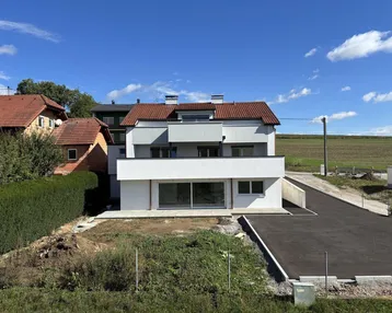 Modernes Einfamilienhaus mit weißer Fassade, rotem Ziegeldach und neu angelegter Zufahrt in ländlicher Umgebung.