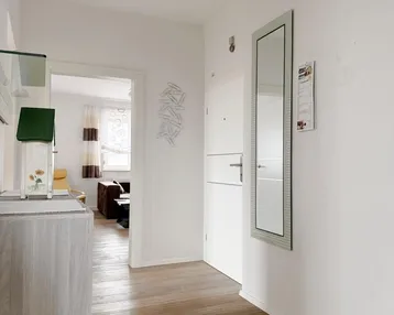 Heller Flur mit Holzoptik-Boden und einem Spiegel, der in den Wohnbereich führt.