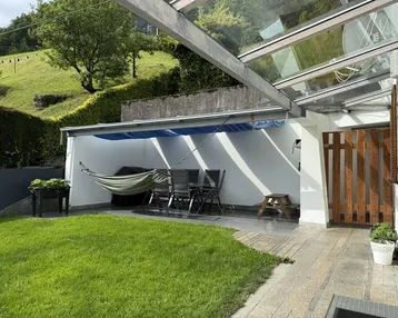 Überdachte Terrasse mit Gartenmöbeln und Hängematte, umgeben von Grünflächen und Hügeln.