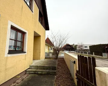 Seitenansicht des Hauses mit gepflastertem Weg und Holzzaun unter bewölktem Himmel.