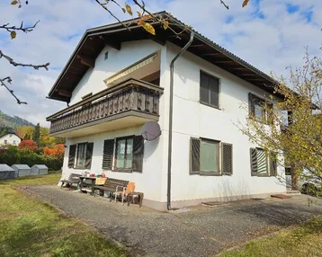Weißes Einfamilienhaus mit Holzbalkon, Fensterläden und kleinem Sitzbereich im Freien.